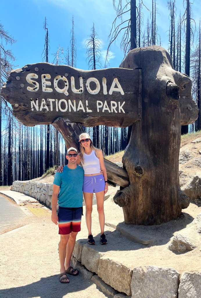 An outdoorsy couple poses in front of the Sequoia national park sign. They are both wearing shorts, hats, and sunglasses. 
